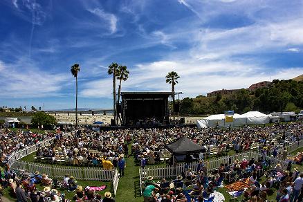 Blue sky hanging over an outdoor concert on an open field filled with people on a bright sunny day with a beach in the background 